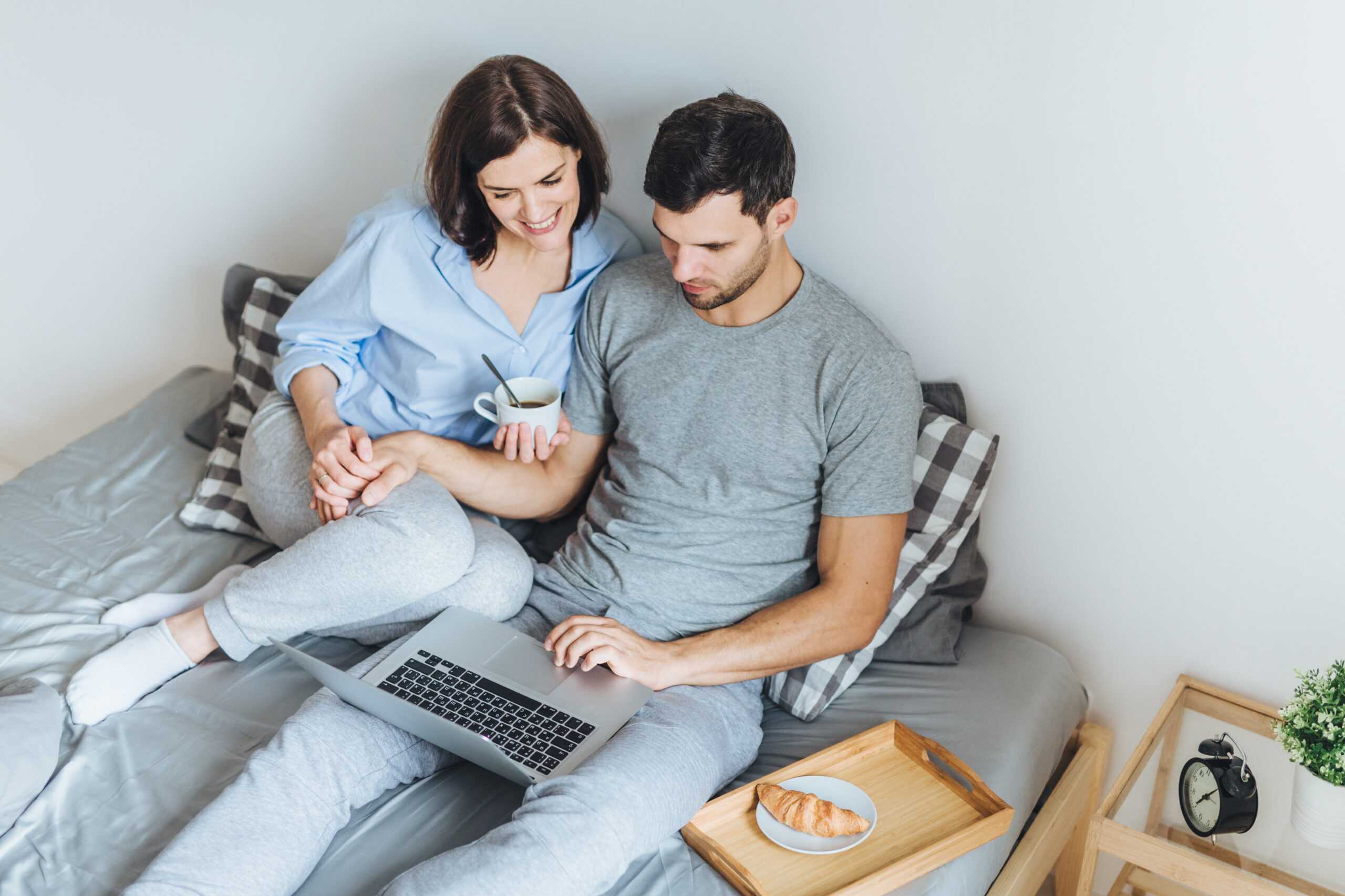 Wife and husband make shopping online, choose present for their little child in internet, look at laptop computer, enjoy togetherness in bedroom, drink hot beverage. People, leisure, technology