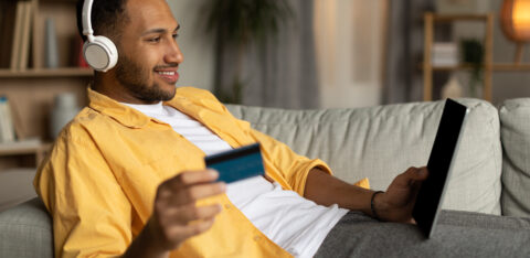 Cheerful man with laptop and credit card lying on couch, wearing headphones, shopping online at home