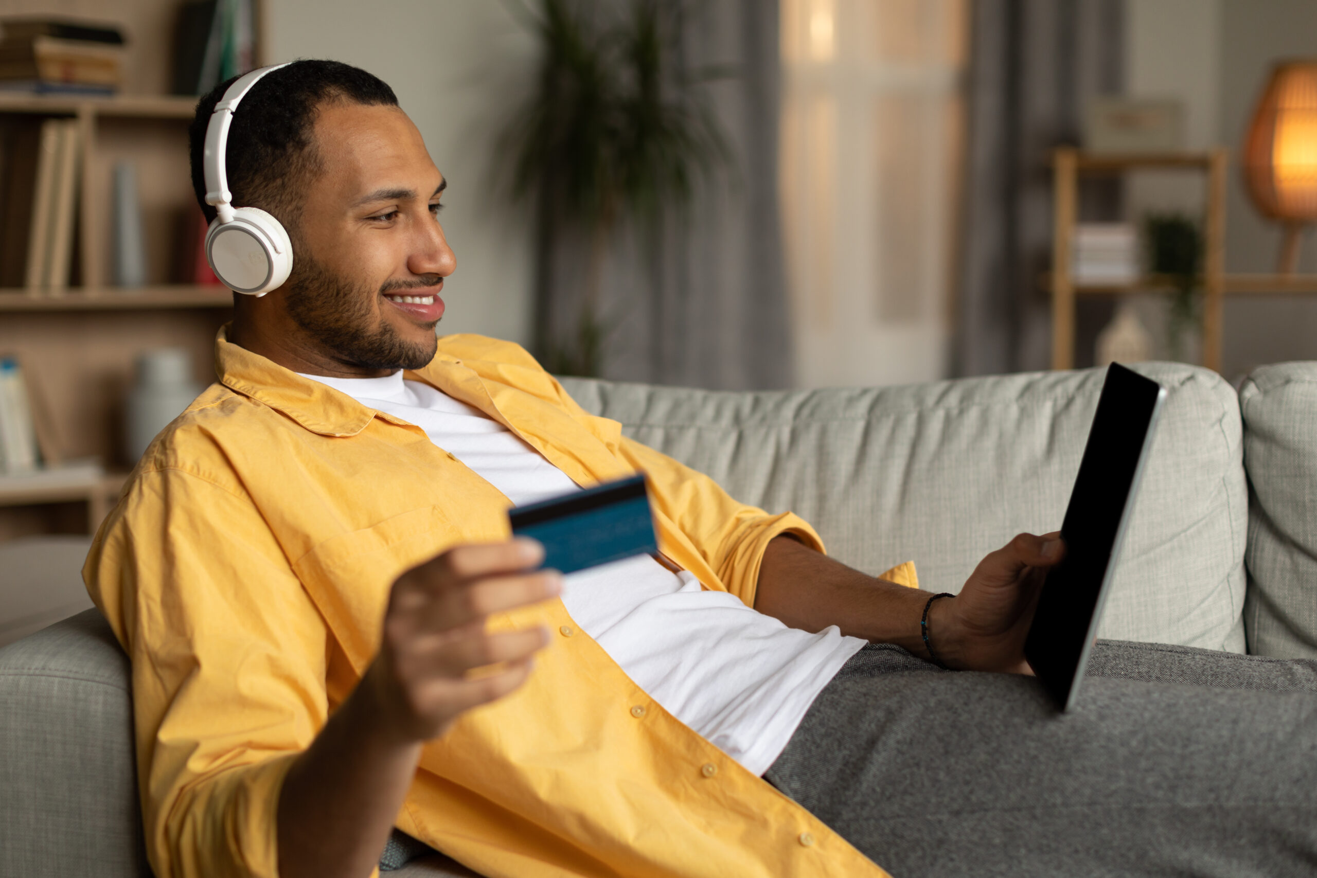 Cheerful man with laptop and credit card lying on couch, wearing headphones, shopping online at home