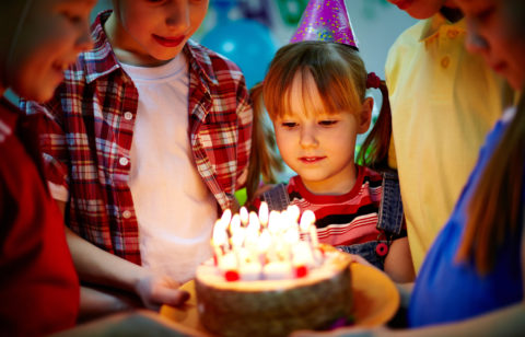 A small circle of children holding up a birthday cake for one to blow out the candles.