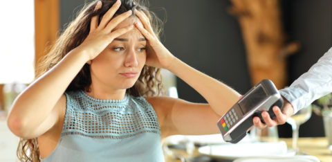 A worried person throws their hands on her head as a restaurant server extends a payment terminal toward them to pay the bill.