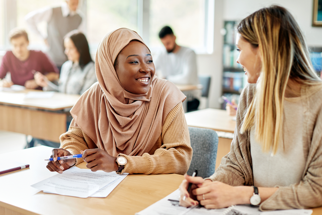 Happy Muslim student talking to her classmate both sitting at a desk