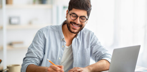 Happy arab man writing business report on laptop and taking notes, sitting at workplace at home interior