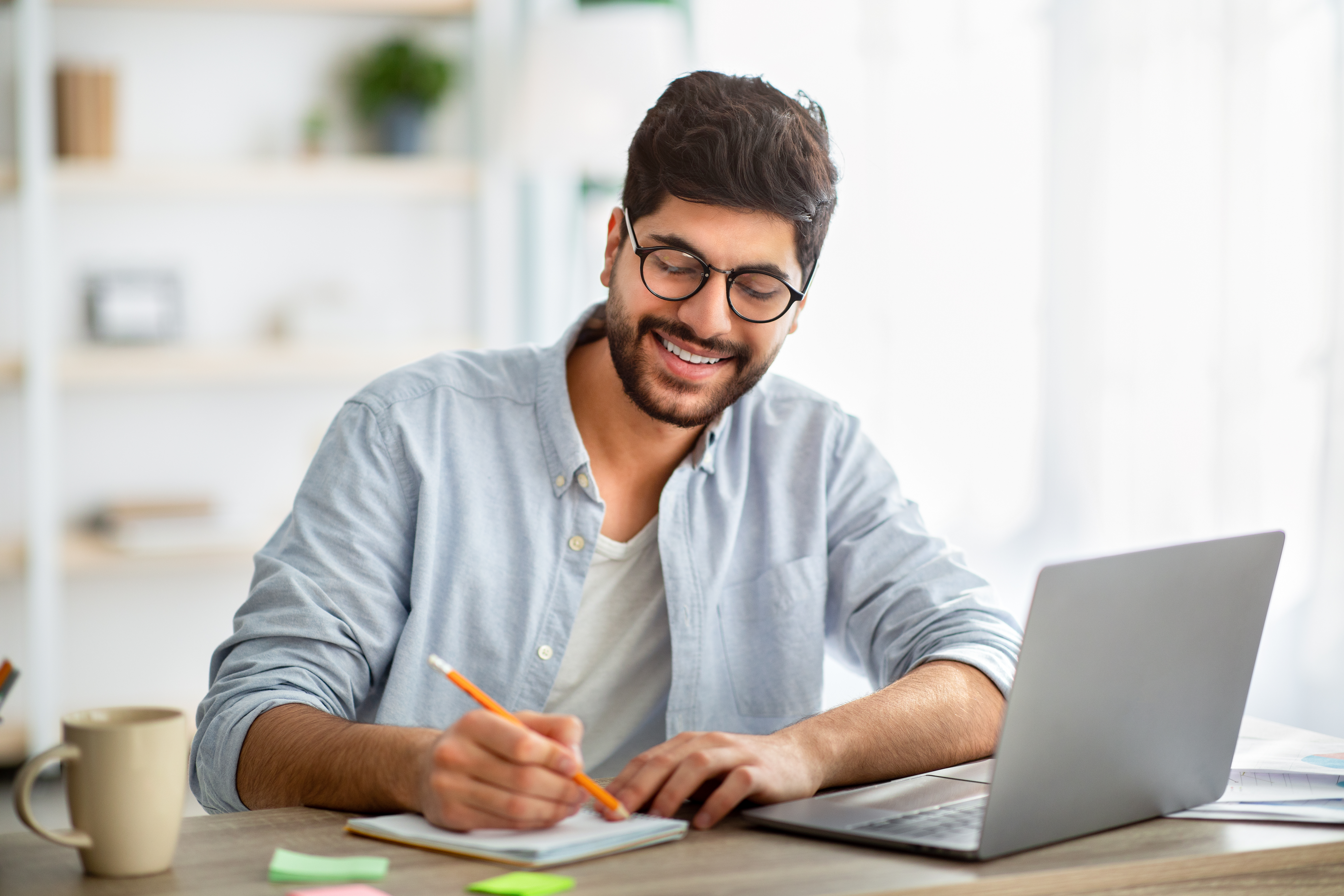 Happy arab man writing business report on laptop and taking notes, sitting at workplace at home interior